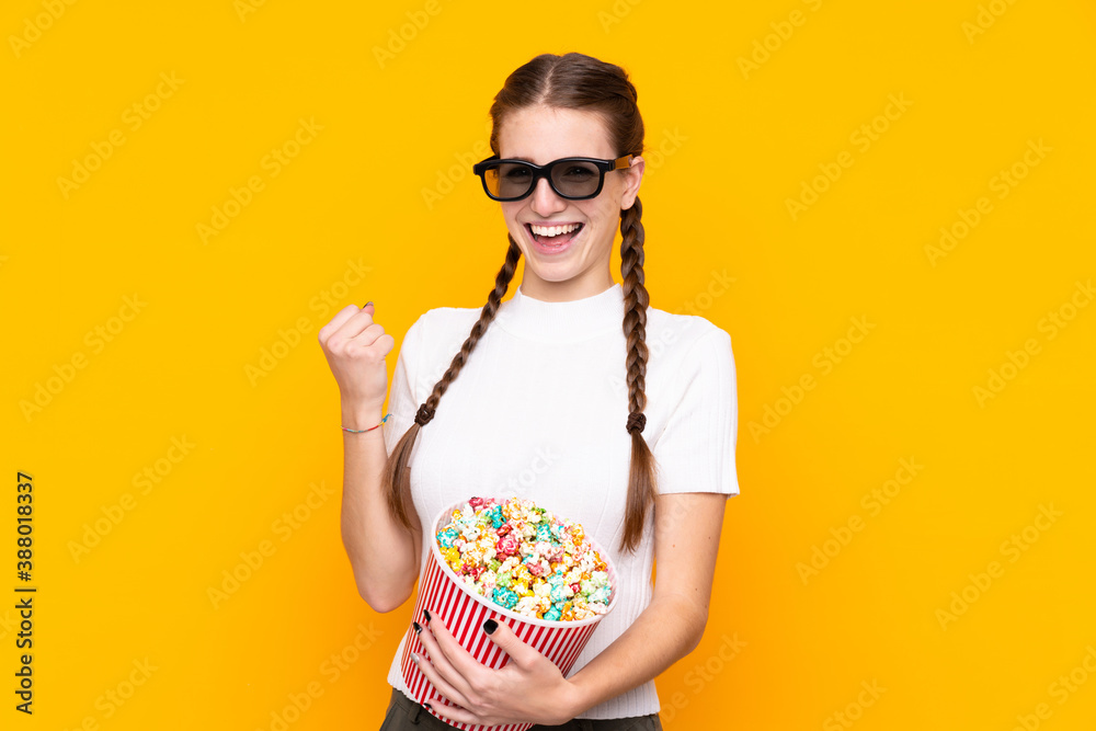Obraz premium Young woman over isolated yellow background with 3d glasses and holding a big bucket of popcorns