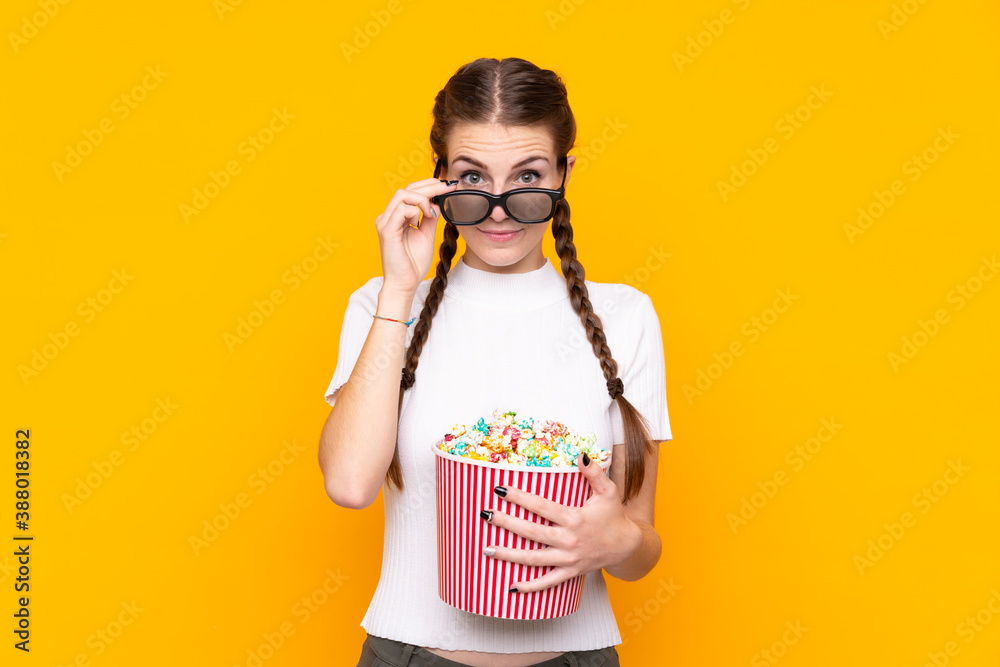 Young woman over isolated yellow background surprised with 3d glasses and holding a big bucket of popcorns