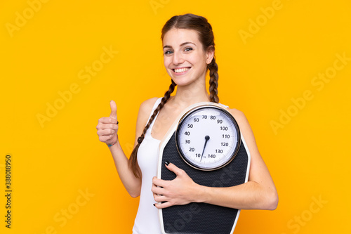 Young woman over isolated yellow background holding weighing machine with thumb up