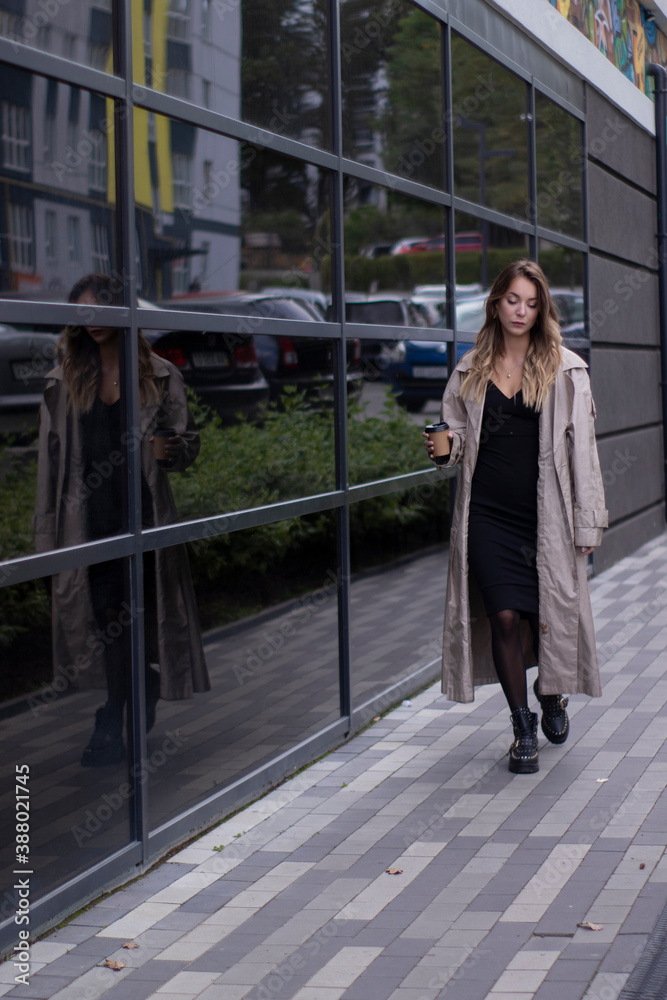 Fototapeta premium beautiful slim young brunette in fashionable trench coat and black dress walking through the street. autumn, yellow leaves