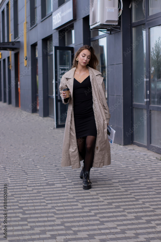 beautiful slim young brunette in fashionable trench coat and black dress walking through the street. autumn, yellow leaves