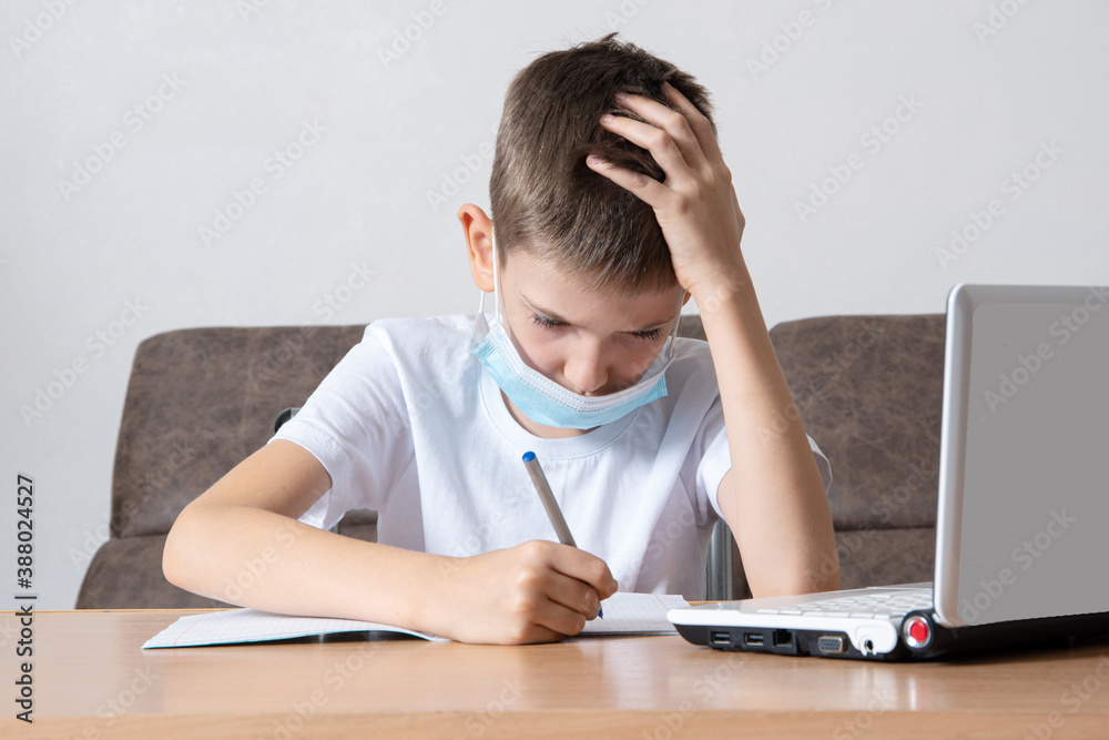 A smart school boy writes information into his notebook while sitting at his desk in front of a laptop. Online education A smart school boy writes information into his notebook while sitting at his desk in front of a laptop. Online education