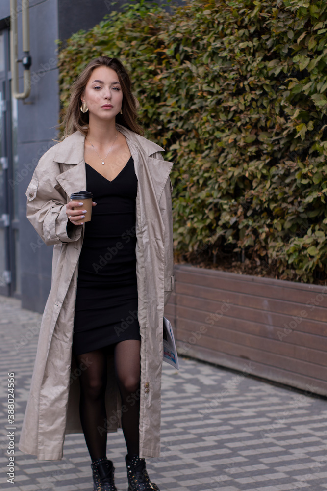 Fototapeta premium beautiful slim young brunette in fashionable trench coat and black dress walking through the street. autumn, yellow leaves