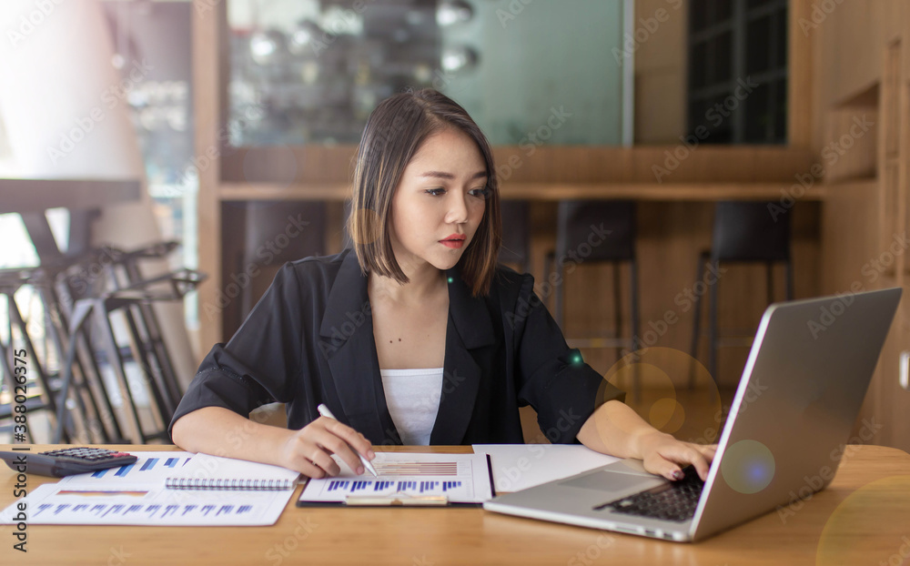 A beautiful young businesswoman sits on a desk using a laptop and checks the graph table to calculate the financial statements within her work station.