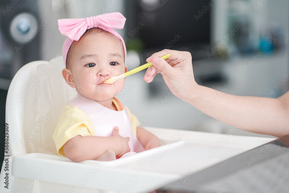 10-month old asian baby girl in a pink bib and hair bow sitting on a white high chair. Her mother giving healthy food by spoon in the morning at home 10-month old asian baby girl in a pink bib and hair bow sitting on a white high chair. Her mother giving healthy food by spoon in the morning at home