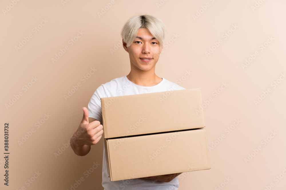 Young asian man over isolated background holding a box to move it to another site with thumb up Young asian man over isolated background holding a box to move it to another site with thumb up