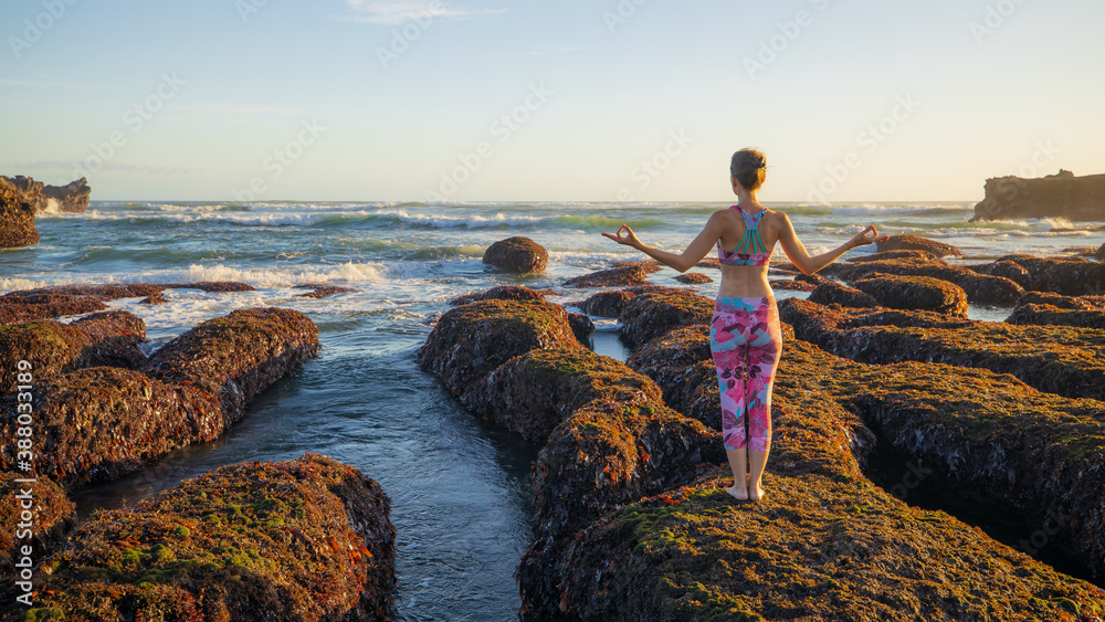 Young Caucasian woman standing on big stone during low tide. Yoga practice at the beach. Hands in gyan mudra. Bali. View from back.
