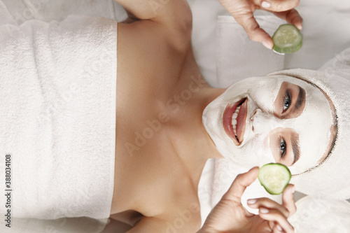 Beautiful young woman is getting facial clay mask at spa, lying with cucumbers on eyes. skin care.