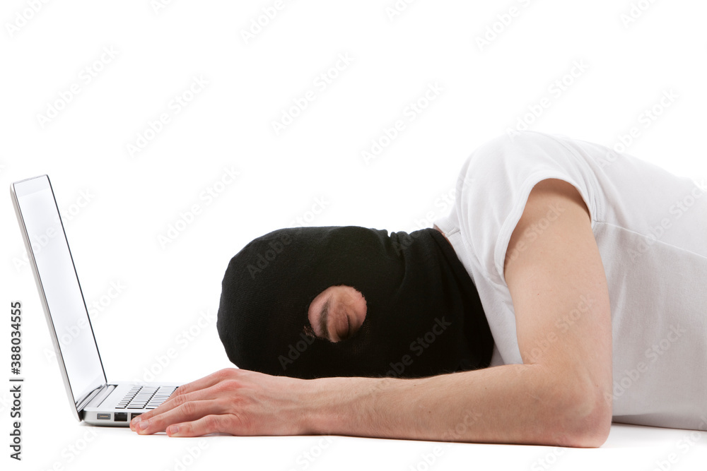 Naklejka premium Tired young caucasian man with black balaclava helmet hacking a laptop. Studio photo.