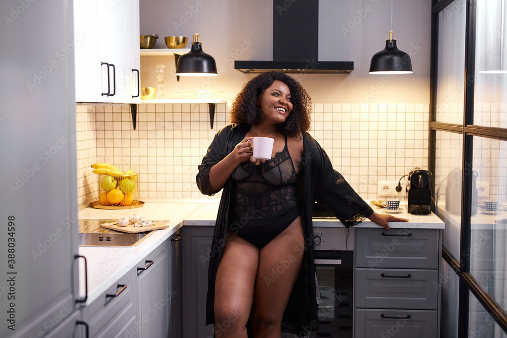beautiful black overweight lady drinking tea at home in the morning, happy day begins from hot cup of tea or coffee, female stands in underwear, half naked