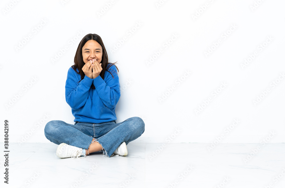 Young mixed race woman sitting on the floor isolated on white background happy and smiling covering mouth with hands