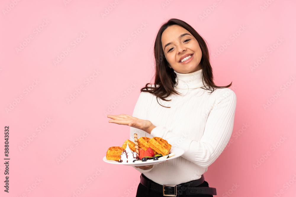 Young brunette woman holding waffles over isolated pink background presenting an idea while looking smiling towards