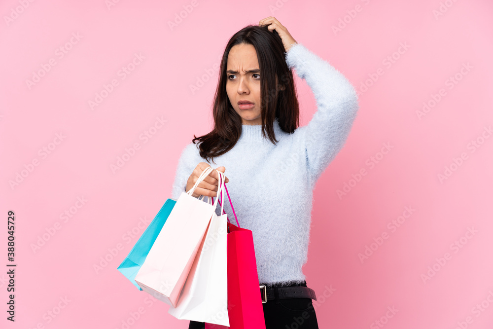Young woman with shopping bag over isolated pink background having doubts while scratching head Young woman with shopping bag over isolated pink background having doubts while scratching head