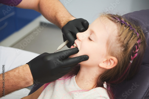 Portrait of little girl has a dental examination while sitting at dentist office. Male dentist is checking the young patient's mouth.