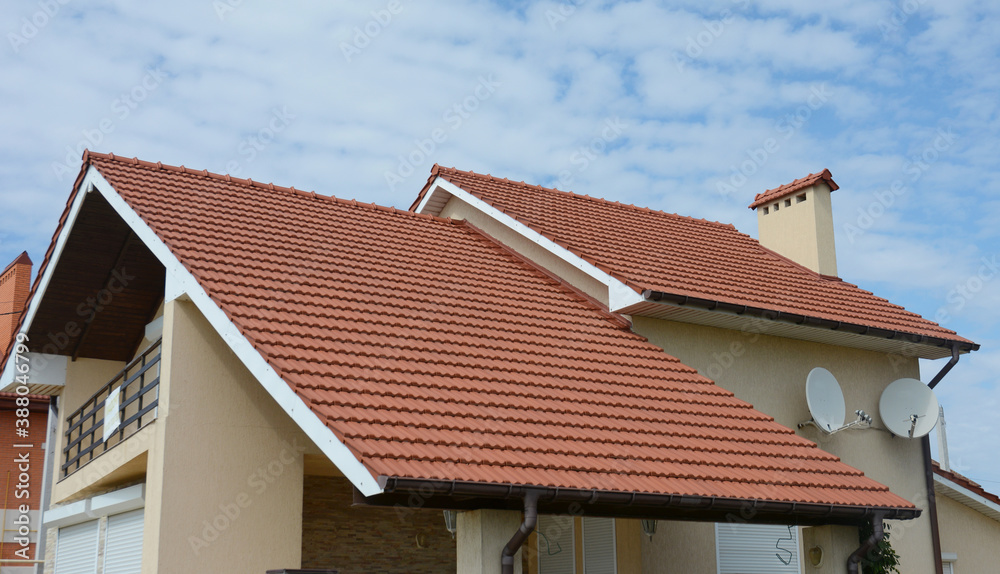 A close-up on a stucco house with a red metal tiled double gabled roof, a chimney, fascia boards, rain gutters, two satellite dish antennas and an attic open balcony against blue sky.