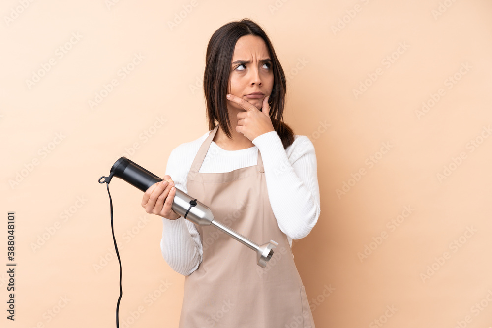 Young brunette girl using hand blender over isolated background having doubts and with confuse face expression