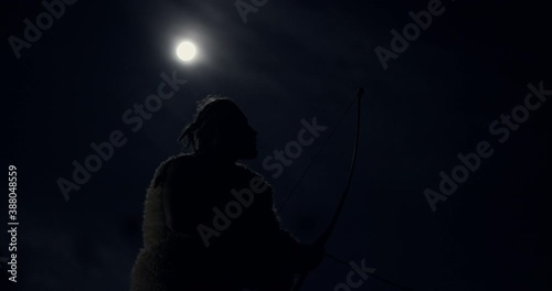 Primitive man hunting at night in full moon. From below side view of primeval male with bow and arrows hunting at night in nature on background of dark sky with full moon