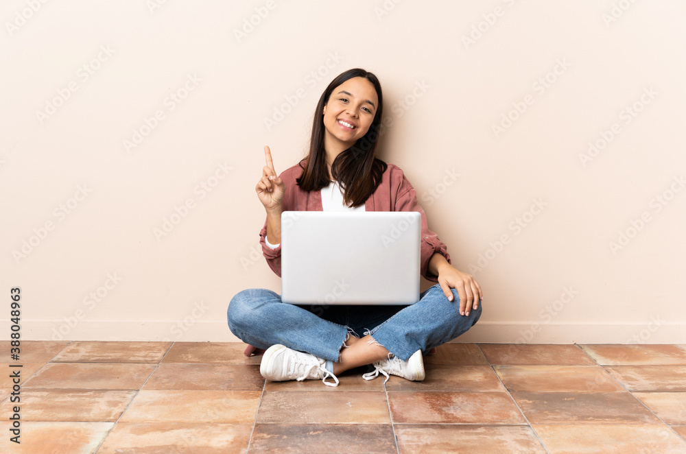 Young mixed race woman with a laptop sitting on the floor showing and lifting a finger in sign of the best Young mixed race woman with a laptop sitting on the floor showing and lifting a finger in sign of the best