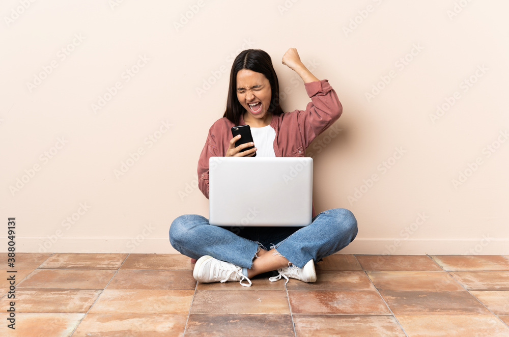 Young mixed race woman with a laptop sitting on the floor with phone in victory position Young mixed race woman with a laptop sitting on the floor with phone in victory position