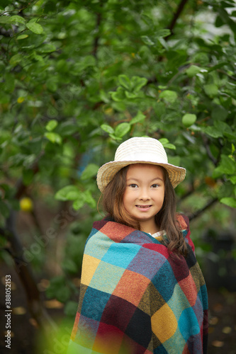 portrait of adorable asian little girl around plants, child is wrapped in plaid, wearing hat, look at camera. in the garden