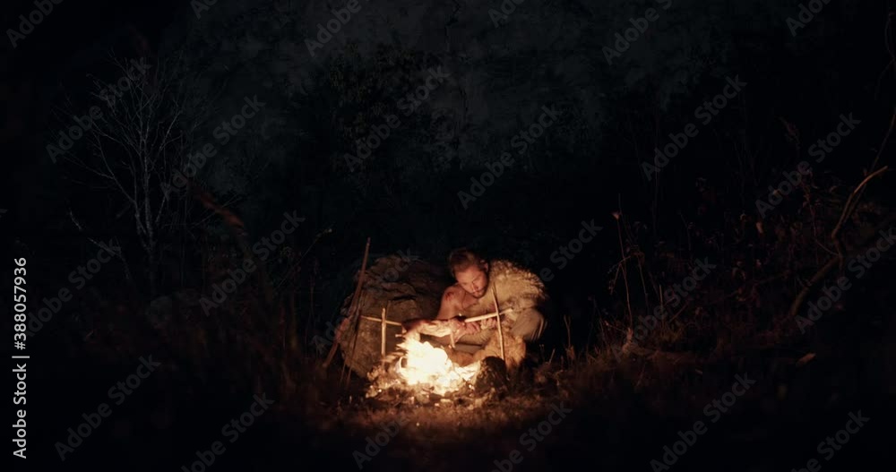 Savage man roasting meat over bonfire. Lonely savage male hunter sitting in front of burning bonfire with killed animal roasted over flame in dark forest at night