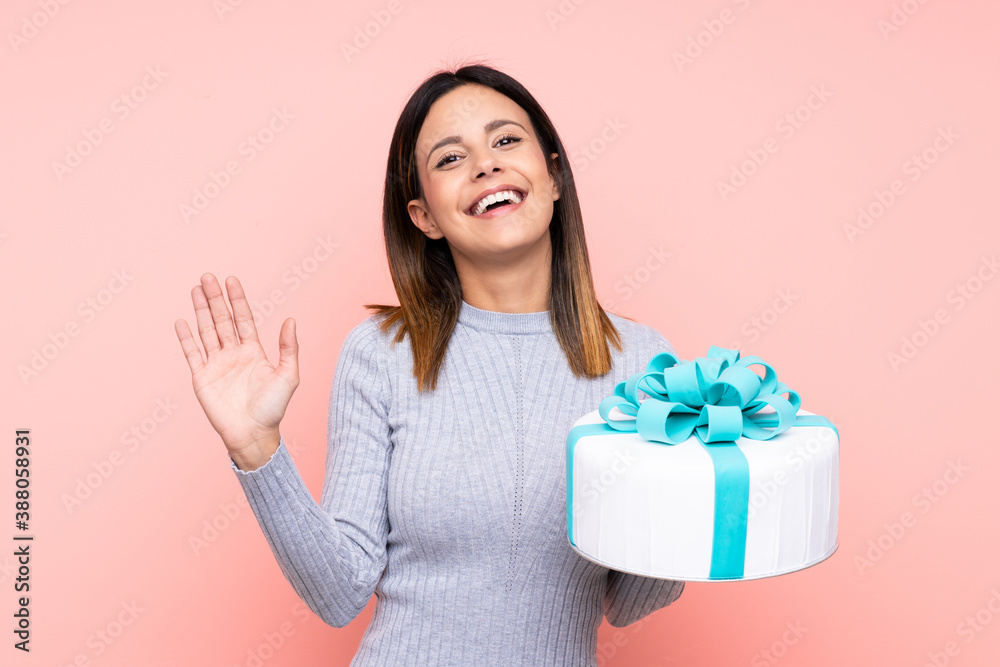 Woman holding a big cake over isolated pink background saluting with hand with happy expression Woman holding a big cake over isolated pink background saluting with hand with happy expression
