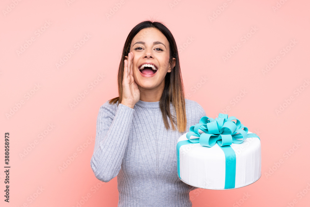 Woman holding a big cake over isolated pink background shouting with mouth wide open Woman holding a big cake over isolated pink background shouting with mouth wide open