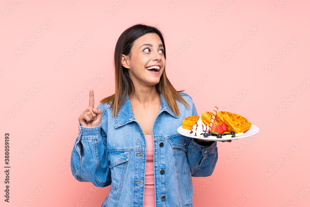 Brunette woman holding waffles over isolated pink background intending to realizes the solution while lifting a finger up Brunette woman holding waffles over isolated pink background intending to realizes the solution while lifting a finger up