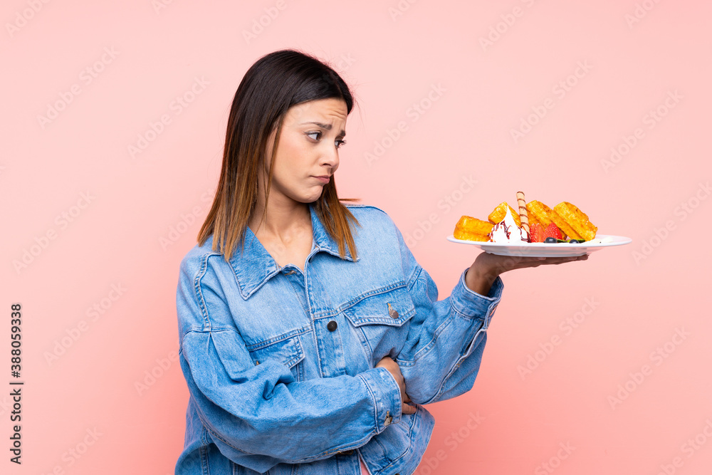 Brunette woman holding waffles over isolated pink background with sad expression Brunette woman holding waffles over isolated pink background with sad expression
