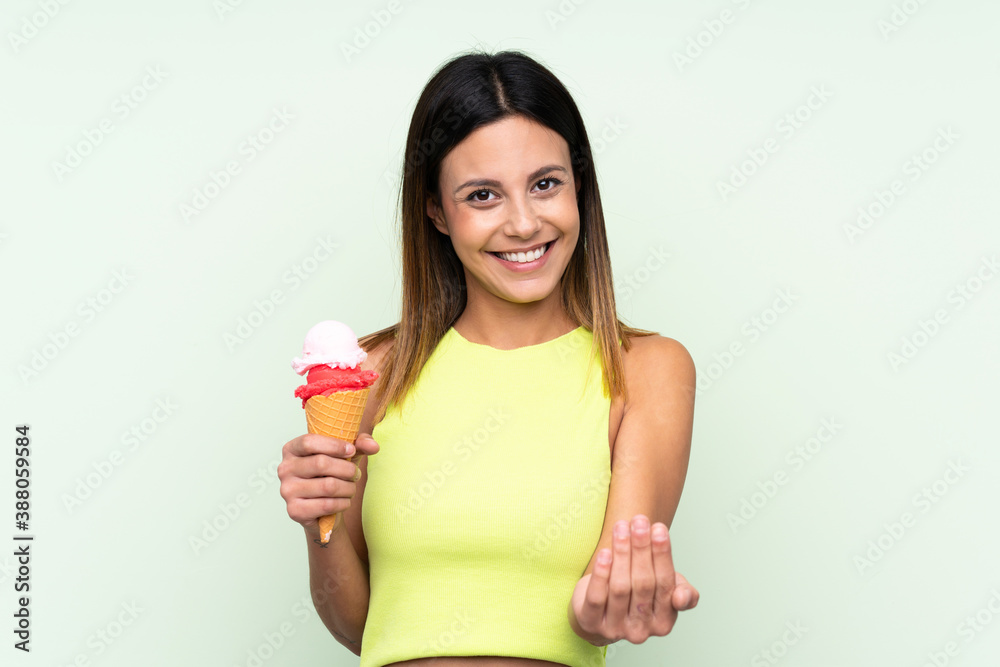 Brunette woman holding a cornet ice cream over isolated green background inviting to come with hand. Happy that you came Brunette woman holding a cornet ice cream over isolated green background inviting to come with hand. Happy that you came