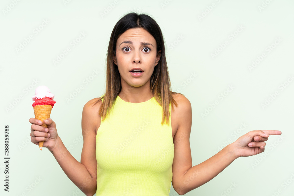 Brunette woman holding a cornet ice cream over isolated green background surprised and pointing side Brunette woman holding a cornet ice cream over isolated green background surprised and pointing side