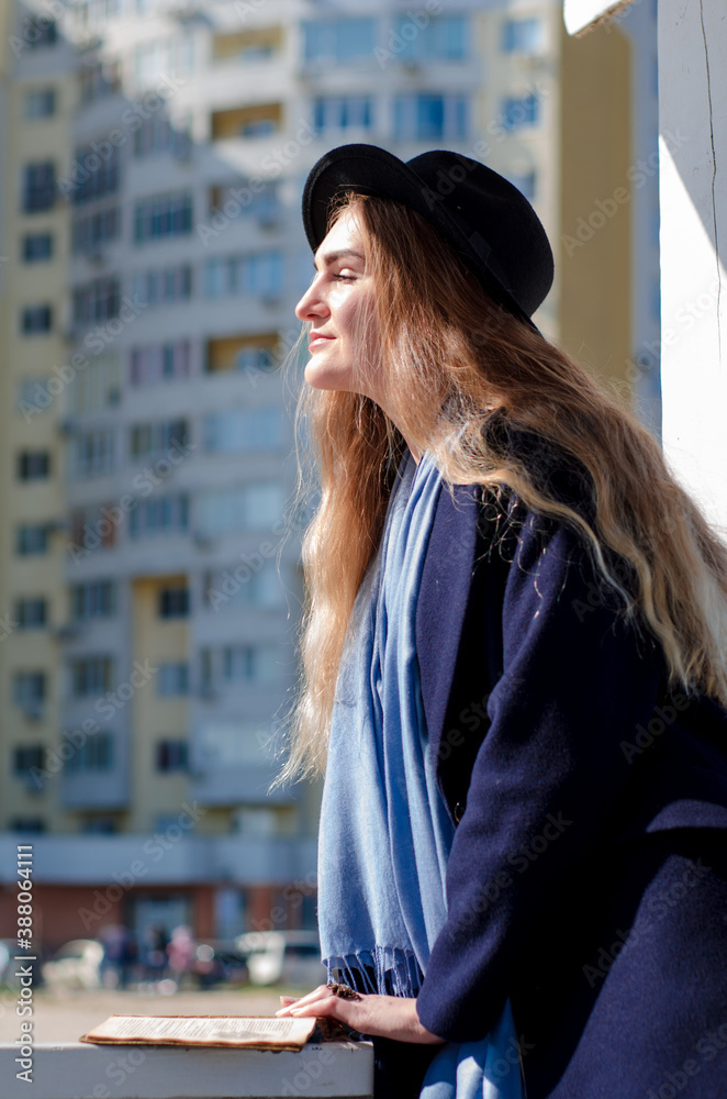 Young caucasian happy smiling girl, in coat and hat in spring city, under rays of spring sun, in background of building Young caucasian happy smiling girl, in coat and hat in spring city, under rays of spring sun, in background of building