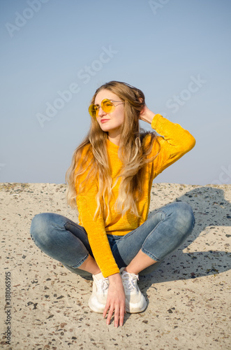 Beautiful blonde girl in yellow sweater and jeans, sneakers sits on concrete surface against blue sky in yellow glasses