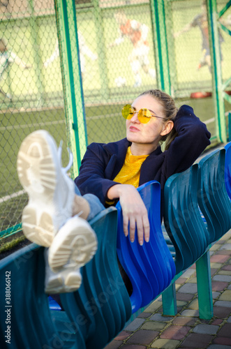 Beautiful blonde girl in yellow sweater, white sneakers and blue coat sits on seats for spectators on sports field