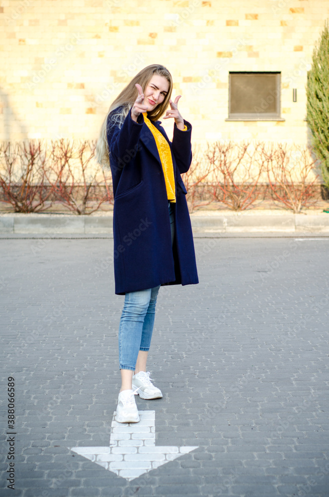 Fototapeta premium Beautiful young blonde girl in white sneakers and blue coat, having fun, laughing in empty parking lot, near white arrow