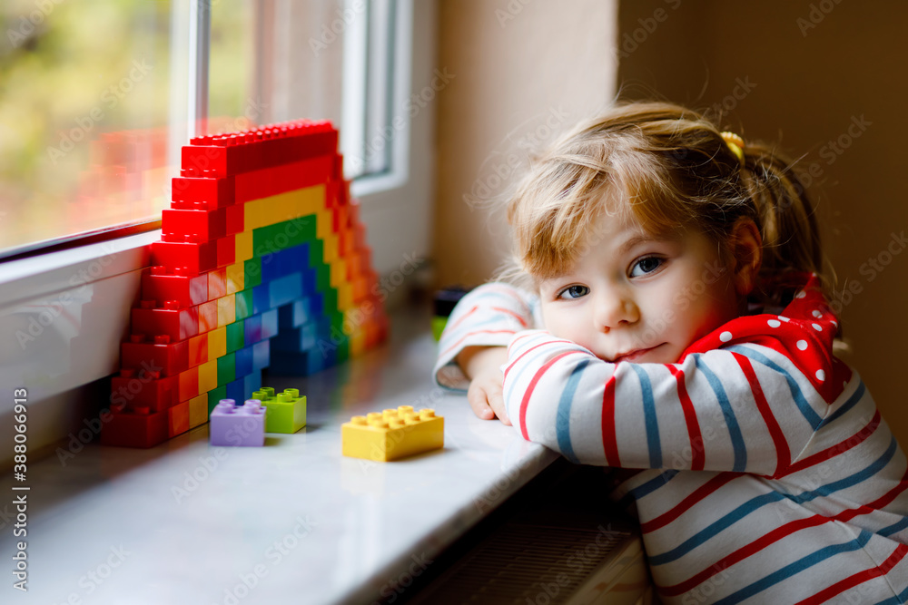 Cute little toddler girl by window create rainbow with colorful plastic blocks during pandemic coronavirus quarantine. Children made and paint rainbows around the world as sign. Cute little toddler girl by window create rainbow with colorful plastic blocks during pandemic coronavirus quarantine. Children made and paint rainbows around the world as sign.