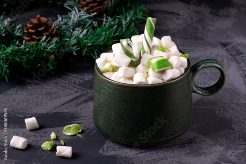 Hot chocolate with marshmallows topped with mint candies and candy canes in a green mug on the grey table. Festive warming drink