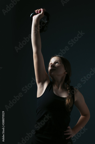 the teenage girl holding the kettlebell in the air with the arm on her heep and looking at it over the dark background © 90783759