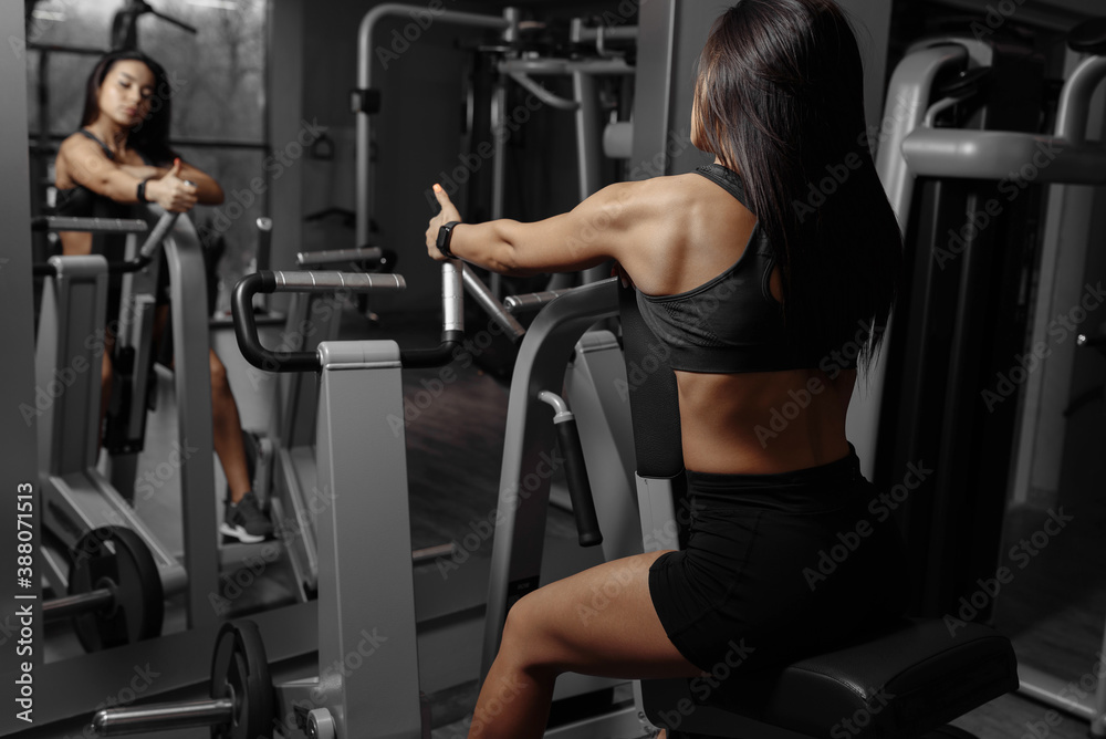 Fototapeta premium Back view of strong muscular sporty woman who workout on exercise machine in front of the mirror in fitness gym