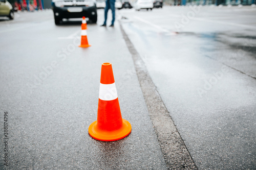 Orange traffic cones with white stripe in driving school on wet asphalt after rain. Blurred gray car on background, male instructor standing near. Starting a driver's license test.