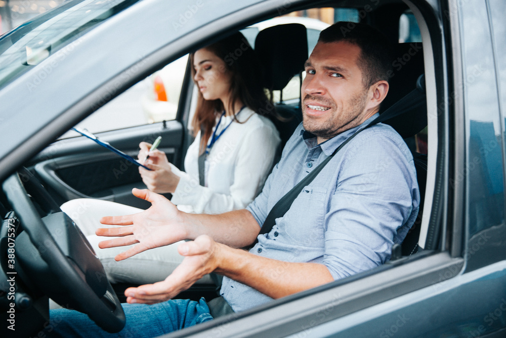 Male student failed exam in driving school, he shrugs up and looks at camera. Man doesn't understand what he is doing wrong. Female auto instructor evaluating male student. Driving school concept. Male student failed exam in driving school, he shrugs up and looks at camera. Man doesn't understand what he is doing wrong. Female auto instructor evaluating male student. Driving school concept.