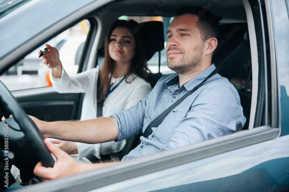 Female driving instructor pointing at object on road by her hand. Diligent male student driving the car and listening what his instructor is saying. Two people in car. Driving lessons concept.