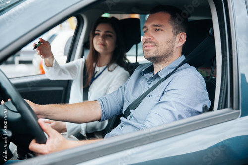 Female driving instructor pointing at object on road by her hand. Diligent male student driving the car and listening what his instructor is saying. Two people in car. Driving lessons concept.