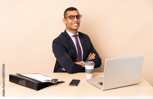 Young business man in his office with a laptop and other documents with arms crossed and happy