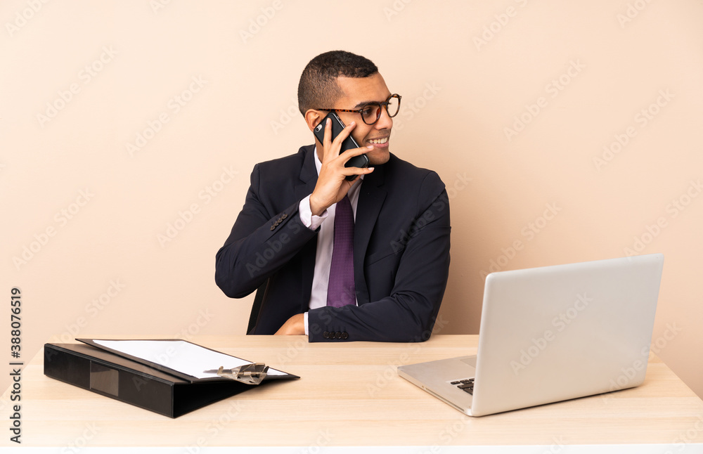 Young business man in his office with a laptop and other documents keeping a conversation with the mobile phone with someone Young business man in his office with a laptop and other documents keeping a conversation with the mobile phone with someone