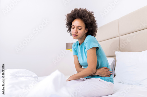 Young woman suffering from abdominal pain while sitting on bed at home. Woman sitting on bed and having stomach ache. Young woman suffering from abdominal pain