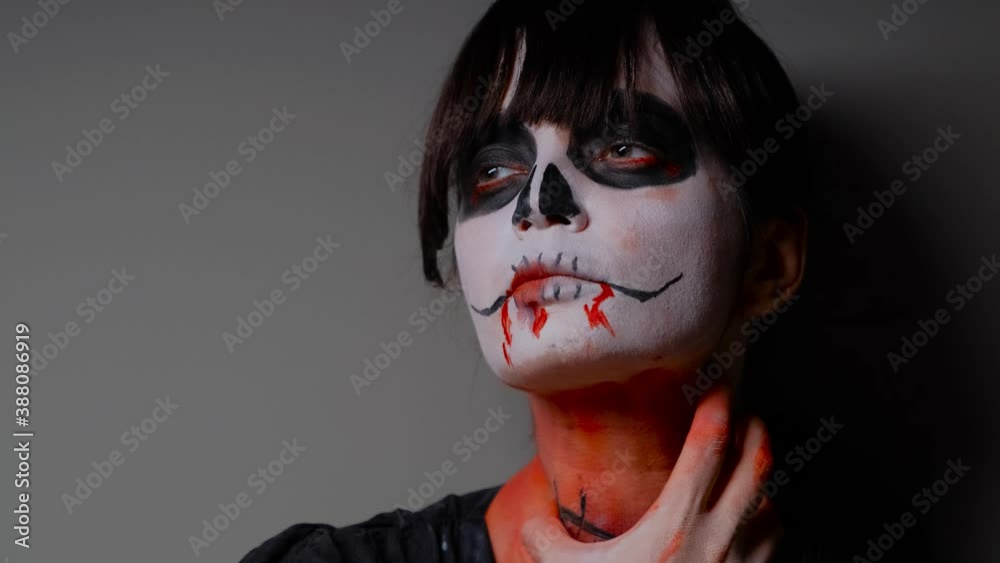A close-up of the face of a zombie girl with dark bangs, who stands against the wall and holds her scarred throat with her painted hand. Halloween Eve. A close-up of the face of a zombie girl with dark bangs, who stands against the wall and holds her scarred throat with her painted hand. Halloween Eve.