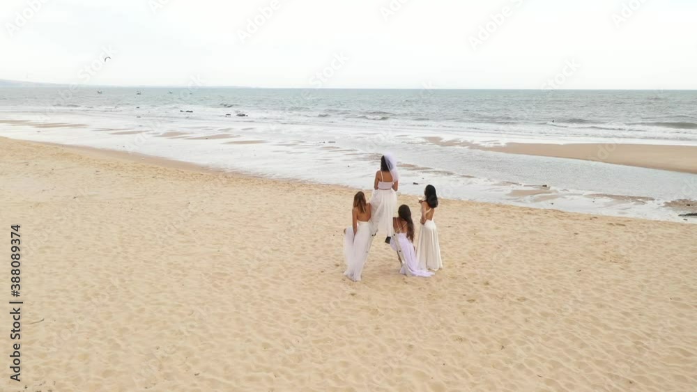 Brides sit on ladder on the beach, aerial view from back to high. Four girls in slow motion video of brides on the beach near the sea from back. Cloudy day and grey water Brides sit on ladder on the beach, aerial view from back to high. Four girls in slow motion video of brides on the beach near the sea from back. Cloudy day and grey water