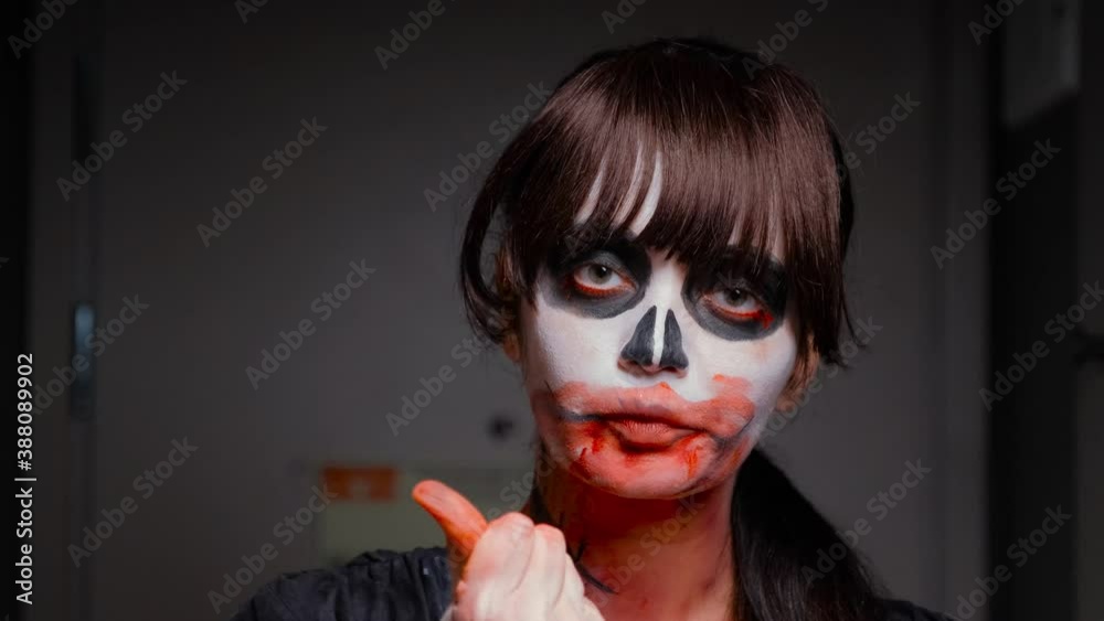 A close-up of the face of a zombie girl with dark bangs, scarred neck and breasts, who smears blood paint on her lips and cheeks. Halloween eve. October holiday.