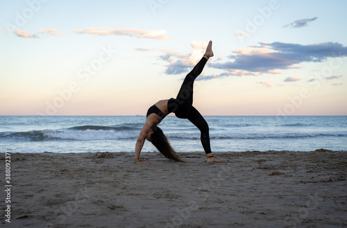 Strong woman practicing yoga on the beach with a beautiful sunset behind her and doing yoga poses.